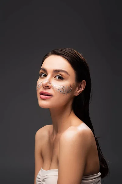 Young brunette woman with bare shoulders and sparkling glitter on cheeks isolated on grey — Stock Photo