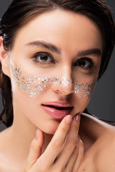 Close up of young brunette woman with sparkling glitter on cheeks touching lips isolated on grey — Stock Photo