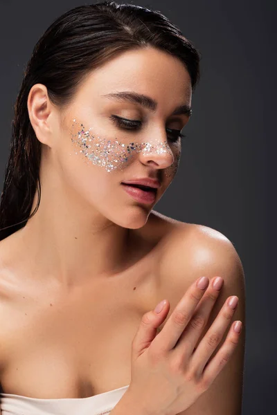 Young woman with sparkling glitter on cheeks touching bare shoulder isolated on grey — Stock Photo