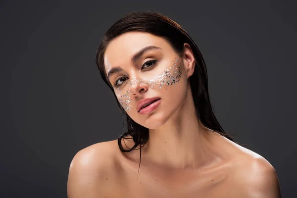 Pretty young woman with bare shoulders and sparkling glitter on cheeks looking at camera isolated on grey — Stock Photo