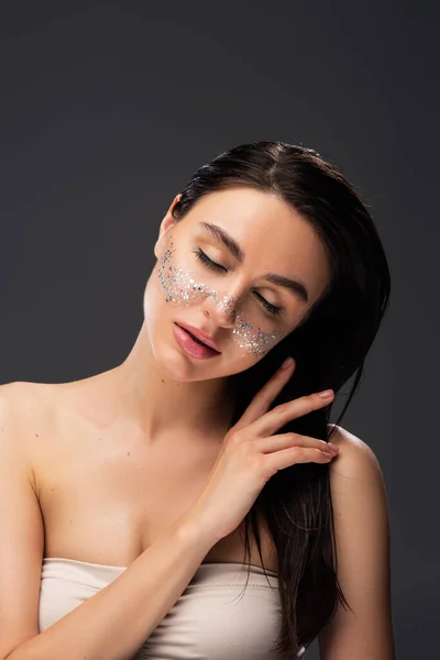 Brunette woman with bare shoulders and sparkling glitter on cheeks touching hair isolated on grey — Stock Photo