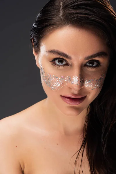 Junge brünette Frau mit natürlichem Make-up und funkelndem Glitzern auf den Wangen, die vereinzelt in die Kamera auf grau blickt — Stockfoto