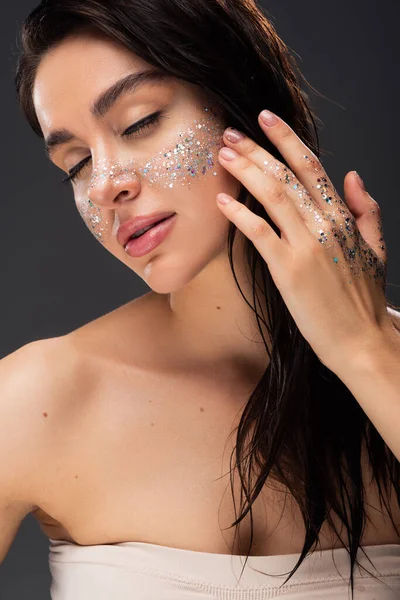 Young brunette woman with closed eyes and sparkling glitter on cheeks and hand isolated on grey — Stock Photo