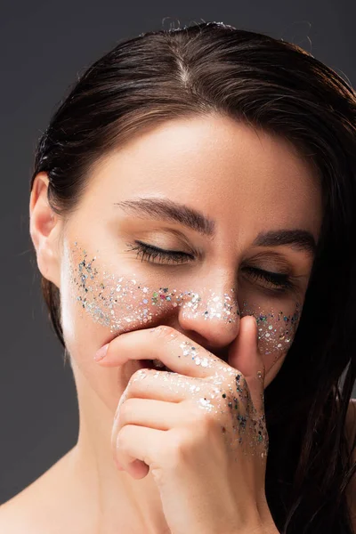 Retrato de una joven morena con brillo brillante en las mejillas riendo y cubriendo la boca aislada en gris - foto de stock