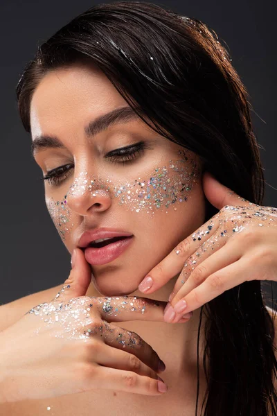 Young brunette woman with sparkling glitter on cheeks and hands touching face isolated on grey — Stock Photo