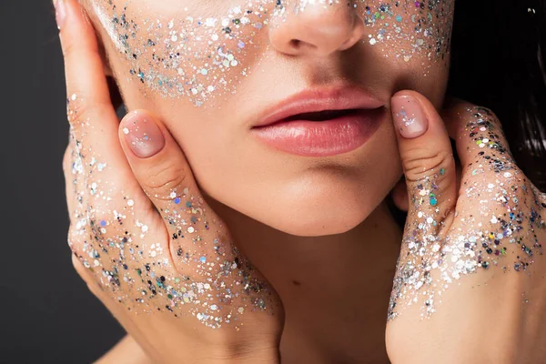 Ausgeschnittene Ansicht einer jungen Frau mit funkelndem Glitzern auf den Wangen und Händen, die Gesicht isoliert auf Grau berühren — Stockfoto