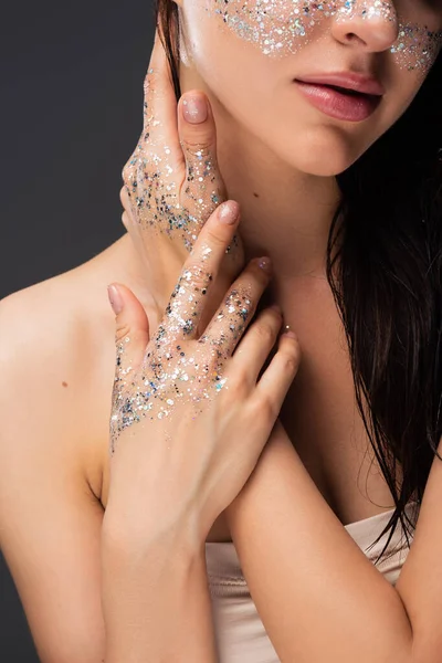 Cropped view of young brunette woman with sparkling glitter on cheeks and hands touching neck isolated on grey — Stock Photo