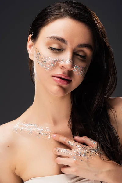 Portrait of young brunette woman with natural makeup and sparkling glitter on cheeks touching chest isolated on grey — Stock Photo