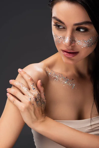 Retrato de mujer con maquillaje natural y brillo brillante en la cara y el cuerpo tocando hombro desnudo aislado en gris - foto de stock