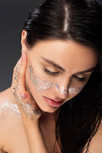 Portrait of woman with natural makeup and sparkling glitter on cheeks and body touching face isolated on grey — Stock Photo