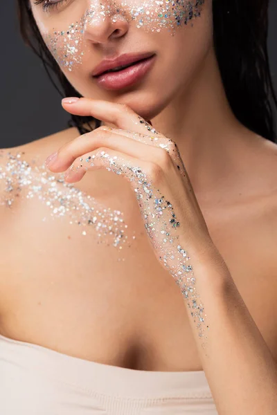 Ausgeschnittene Ansicht einer jungen Frau mit funkelndem Glitzern auf den Wangen und Körperberührung am Kinn isoliert auf grau — Stockfoto