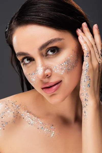 Jeune femme avec des paillettes scintillantes sur les joues et le corps ajuster la publicité capillaire en regardant la caméra isolée sur gris — Photo de stock