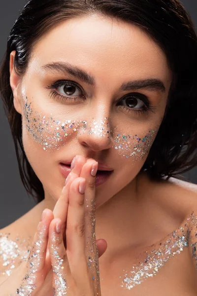 Porträt einer jungen Frau mit Glitzern auf den Wangen und betenden Händen, die isoliert auf grau in die Kamera blickt — Stockfoto