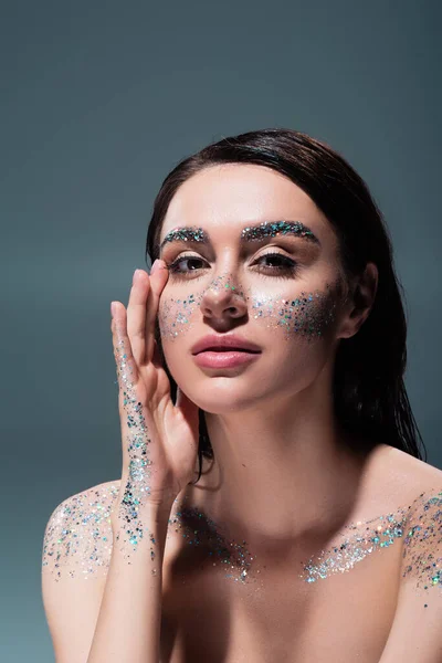 Portrait of brunette woman with glitter on cheeks and body looking at camera isolated on grey — Stock Photo