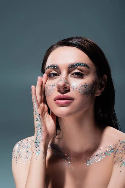 Portrait de femme brune avec des paillettes sur les joues et le corps regardant la caméra isolée sur le gris — Photo de stock