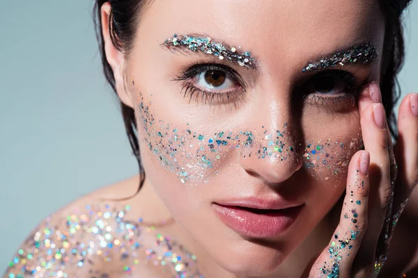 Portrait of brunette woman with glitter on cheeks and eyebrows looking at camera isolated on grey — Stock Photo