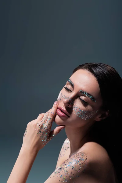 Portrait of young brunette woman with closed eyes and glitter on eyebrows touching face isolated on grey — Stock Photo