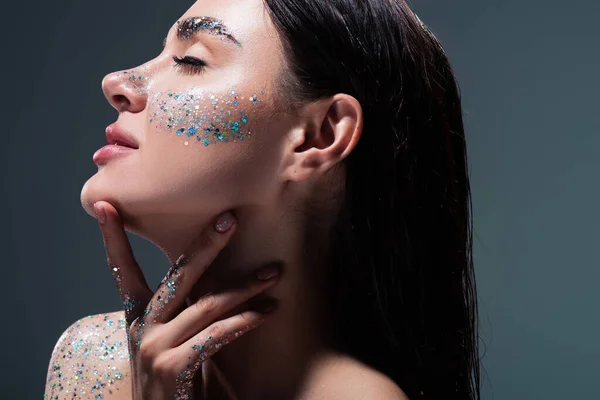 Portrait of young brunette woman with closed eyes and glitter on eyebrows touching neck isolated on grey — Stock Photo