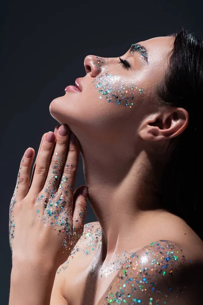 Side view of young woman with closed eyes and glitter on face praying isolated on grey — Stock Photo