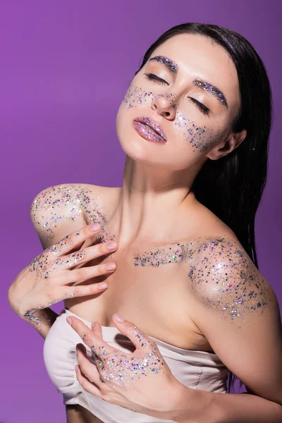 Sensual mujer joven con los ojos cerrados y brillo sobre hombros desnudos aislados en púrpura - foto de stock