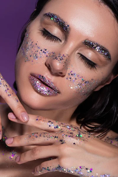 Portrait of young woman with closed eyes and glitter on face isolated on purple — Stock Photo