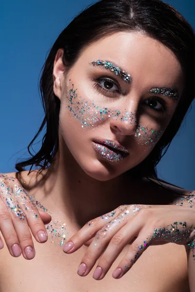 Portrait de jeune femme brune avec des paillettes sur les lèvres et les joues en regardant la caméra isolée sur le bleu — Photo de stock