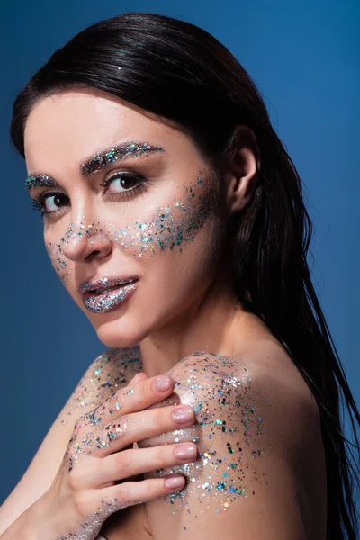 Portrait de jeune femme brune avec des paillettes sur le corps et les joues en regardant la caméra isolée sur le bleu — Photo de stock