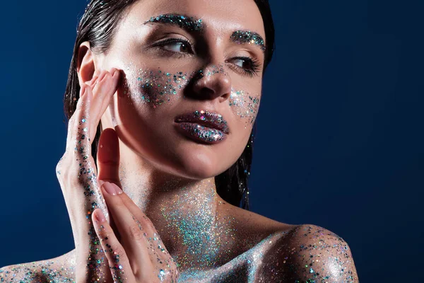 Porträt einer brünetten jungen Frau mit glitzerndem Körper, die Gesicht auf blauem Grund berührt — Stockfoto