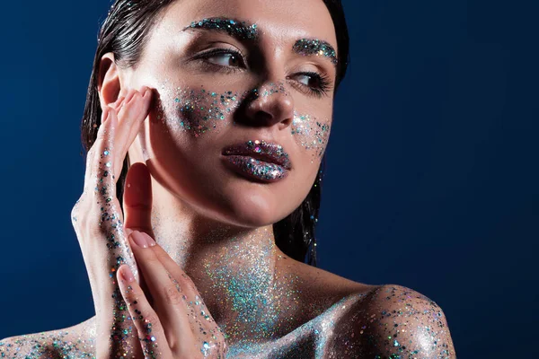 Retrato de morena jovem com brilho no corpo tocando rosto isolado no azul — Fotografia de Stock