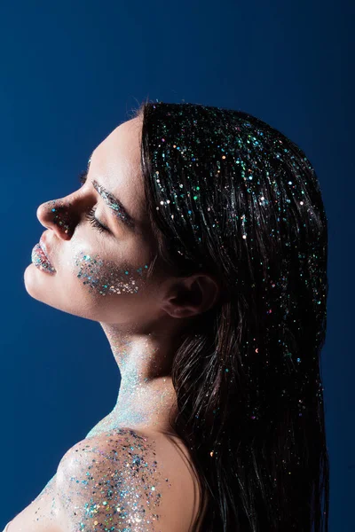 Vista laterale di bruna giovane donna con scintillio scintillante sul corpo e capelli isolati sul blu — Foto stock