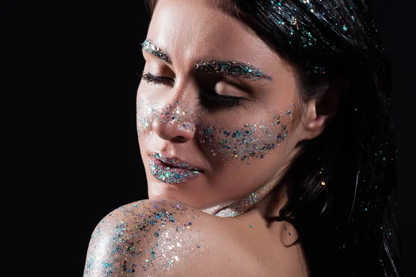 Portrait of young woman with closed eyes and sparking glitter on body isolated on black — Stock Photo