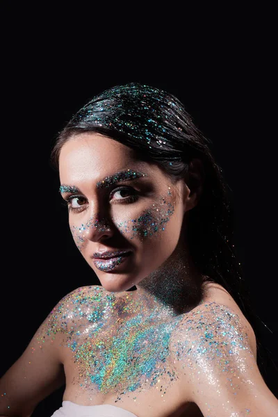 Portrait de jeune femme avec étincelle paillettes sur le corps et le visage en regardant la caméra isolée sur noir — Photo de stock