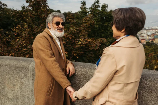 Hombre barbudo y mayor feliz en gafas de sol tomados de la mano con la esposa en gabardina - foto de stock