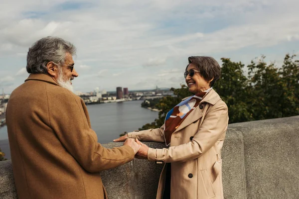 Homme barbu et aîné dans des lunettes de soleil tenant la main avec femme gaie en trench coat — Photo de stock