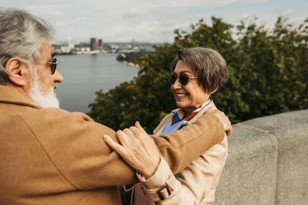 Fröhliche Seniorin mit Sonnenbrille umarmt bärtigen Ehemann im beigen Mantel — Stockfoto