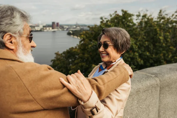 Gaie femme âgée en lunettes de soleil étreignant mari barbu en manteau beige — Photo de stock