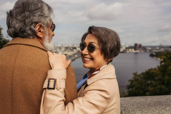 Heureuse femme âgée en lunettes de soleil étreignant mari barbu en manteau beige — Photo de stock