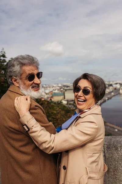 Mujer mayor excitada en gafas de sol con estilo abrazando marido barbudo en abrigo beige - foto de stock