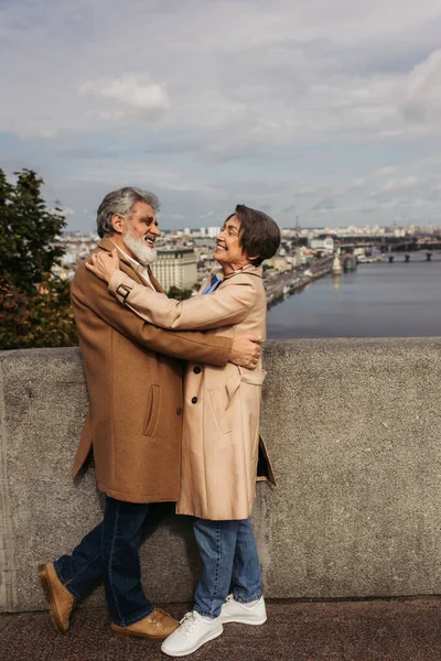 Happy and senior couple in beige coats hugging on bridge near river — Stock Photo