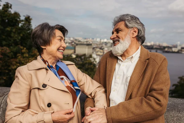 Alegre senior hombre en abrigo mirando sonriente esposa cerca de río en otoño - foto de stock