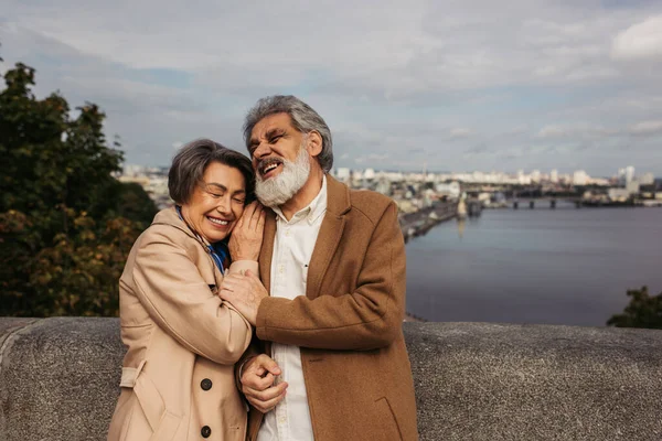 Mulher sênior se apoiando no ombro do marido de casaco e rindo perto do rio — Fotografia de Stock