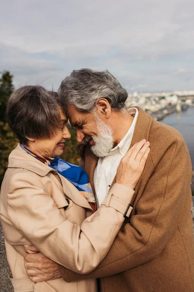 Senior femme étreignant avec mari barbu en manteau et souriant près de rivière — Photo de stock