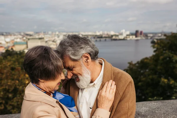 Mujer mayor abrazándose con el marido barbudo con el pelo gris y sonriendo cerca del río - foto de stock