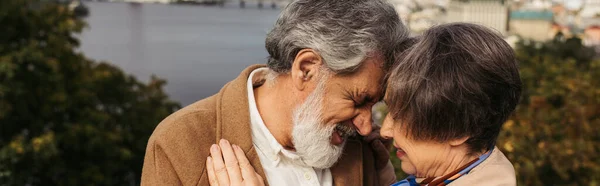 Старша жінка обіймається з бородатим чоловіком з сірим волоссям і посміхається біля річки, банер — стокове фото