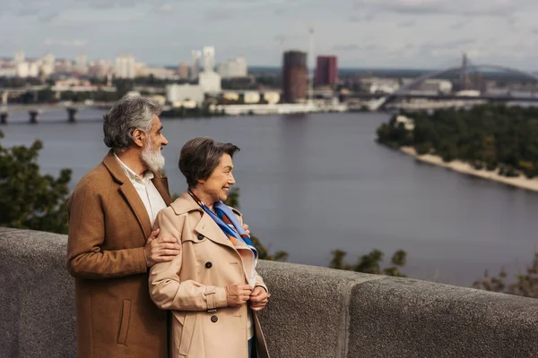 Alegre pareja de ancianos en abrigos beige sonriendo y de pie en el puente cerca del río - foto de stock