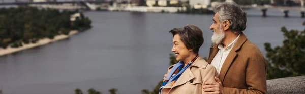 Alegre pareja de ancianos en abrigos beige sonriendo y de pie cerca del río, bandera - foto de stock