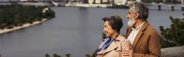 Joyeux couple aîné en manteaux beige souriant et debout près de la rivière, bannière — Photo de stock