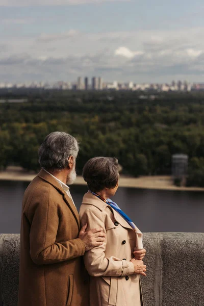 Rückansicht eines älteren Ehepaares in beigen Mänteln, das auf einer Brücke in der Nähe des Flusses steht — Stockfoto