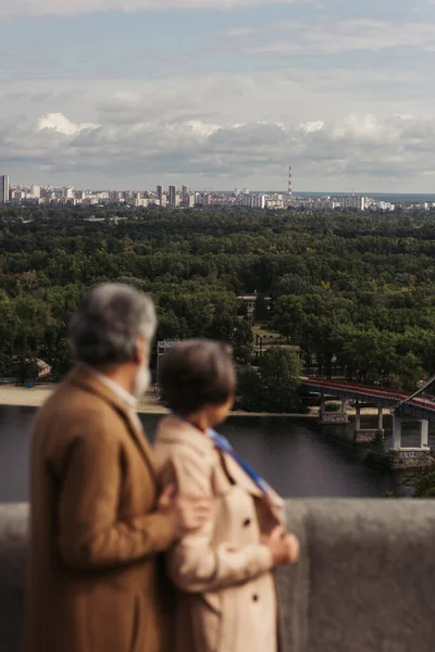 Vue arrière du couple âgé flou en manteaux beiges debout sur un pont près de la rivière — Photo de stock