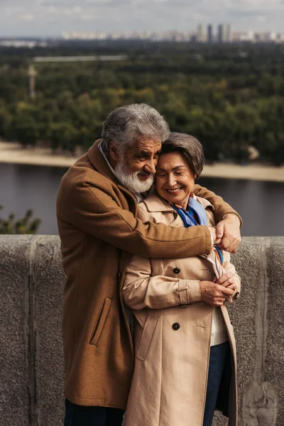 Bearded senior man smiling while hugging joyful wife on bridge near river — Stock Photo